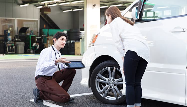 車の前でタブレット端末を用いて説明する整備士の男性や女性客｜タイヤ館